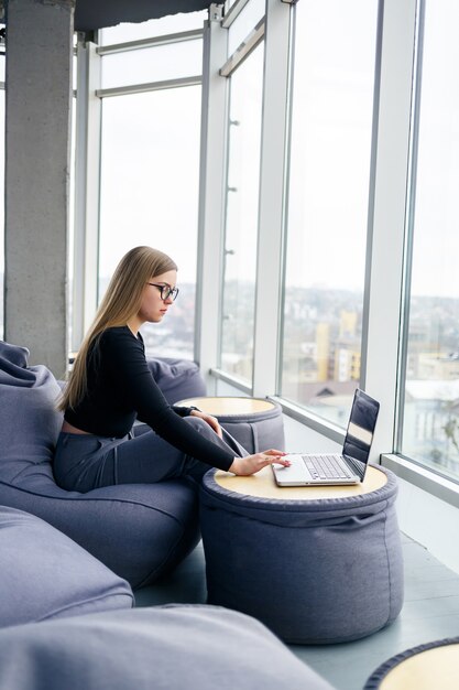 Eine schöne junge Managerin sitzt mit einem Laptop auf einem weichen Sitzpuff in der Nähe des Panoramafensters. Mädchengeschäftsmann, der an einem neuen Projekt arbeitet