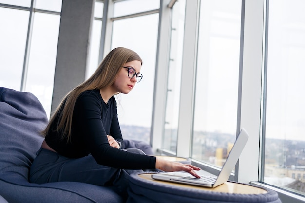 Eine schöne junge Managerin sitzt mit einem Laptop auf einem weichen Sitzpuff in der Nähe des Panoramafensters. Mädchengeschäftsmann, der an einem neuen Projekt arbeitet
