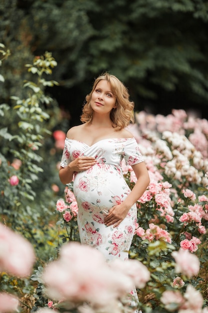 Eine schöne junge schwangere Frau geht in einem Rosengarten. Porträt einer schwangeren Frau in einem Kleid. Sommer.