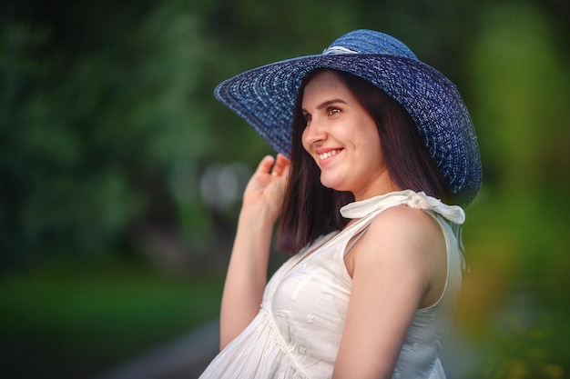 Eine schöne junge schwangere Frau in einem blauen Hut lächelt und schaut weg Gehen Sie im Park und warten Sie auf die Geburt eines Kindes