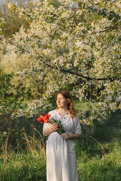 Eine schöne junge schwangere Frau in einem weißen Kleid geht in den Frühlingsgarten