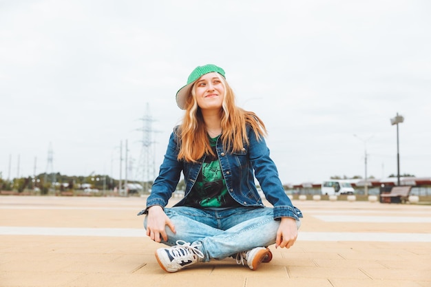 Eine schöne junge Teenagerin in Mütze und Jeansjacke sitzt über einer Skateboard-Generation z