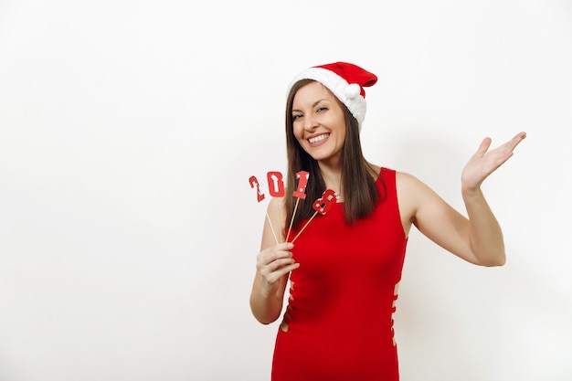 Eine schöne kaukasische junge glückliche Frau mit gesunder Haut und charmantem Lächeln in rotem Kleid und Weihnachtsmütze mit Kartennummer 2018 auf weißem Hintergrund. Sankt-Mädchen isoliert. Neujahrsferienkonzept