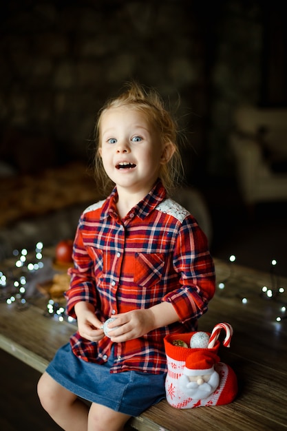 Eine schöne kleine Blondine in einem traditionellen karierten Hemd öffnet einen Schokoriegel von ihrem süßen Weihnachtsgeschenk