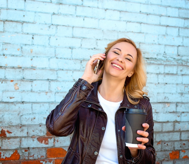 Eine schöne lächelnde blonde Frau mit dem Telefon, das eine Tasse Kaffee zum Mitnehmen hält.