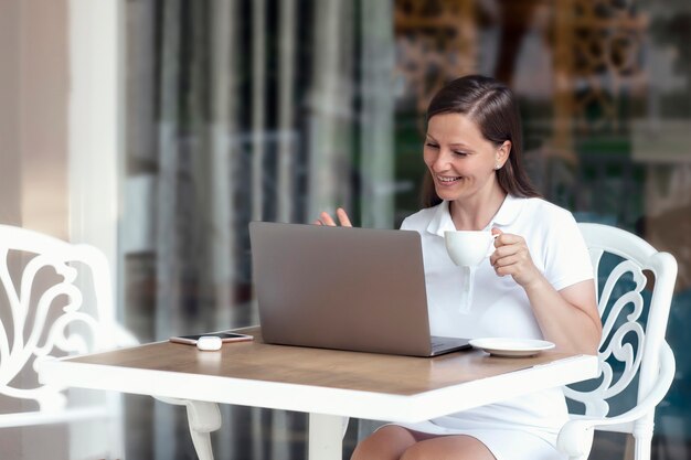 Eine schöne lächelnde Frau in einem weißen Kleid kommuniziert über einen Laptop in einem Café