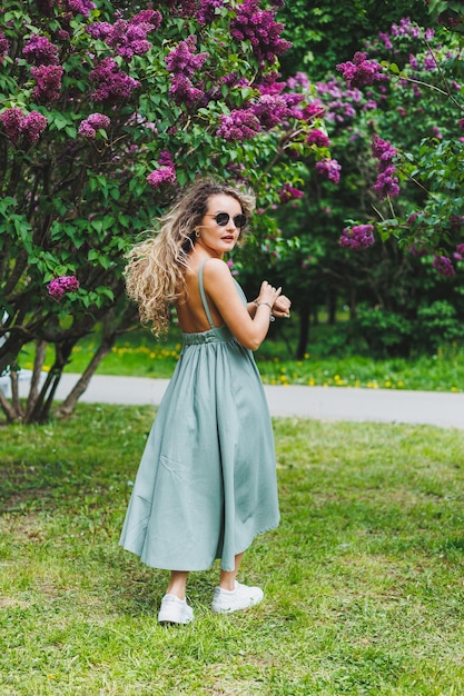 Eine schöne lockige Frau in einem Sommerkleid läuft und freut sich, dass sie eine Sonnenbrille vor dem Hintergrund eines violetten Fliederbusches trägt