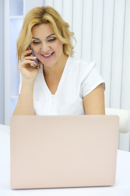 Eine schöne, reife Frau, Kosmetikerin, Kosmetikerin, sitzt in ihrem Büro an einem Tisch mit offenem Laptop. Der Arzt spricht am Telefon und vereinbart einen Termin oder eine Beratung