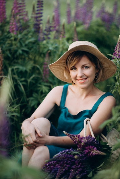 Eine schöne romantische Frau lächelt freudig in einem blauen Kleid und Hut sitzt in einem Feld von lila Lupinenblumen.