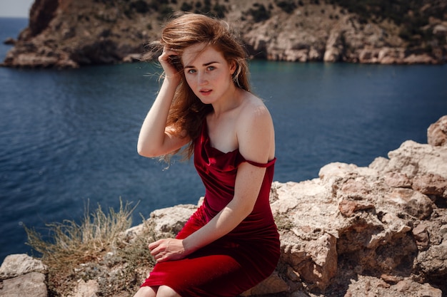 Eine schöne rothaarige Frau im roten Kleid sitzt auf einem Felsen mit herrlichem Blick auf die Seelandschaft. Sommernachmittag Genuss von Freiheit und Einsamkeit