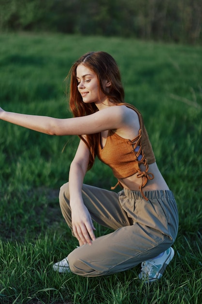 Eine schöne rothaarige Frau sitzt in sportlicher Sommerkleidung im Gras und entspannt sich nach einem Spaziergang in der Natur und blickt auf den Sonnenuntergang