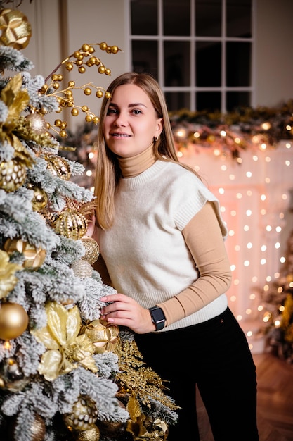 Eine schöne schöne Frau in einem beigen Pullover sitzt auf dem Boden vor dem Hintergrund eines Weihnachtsbaums und schmückt einen Weihnachtsbaum mit Spielzeug in einem geräumigen, mit Girlanden geschmückten Raum.