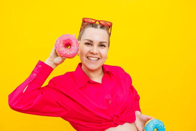 Eine schöne schwangere Frau hält einen süßen Donut in der Hand auf gelbem Hintergrund und erwartet eine Schwangerschaft und Mutterschaft für ein Kind. Das Konzept der gesunden und ungesunden Ernährung Junk Food