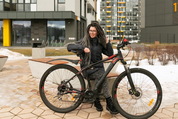 Eine schöne städtische Frau mit Dreadlocks-Frisur, gekleidet in einer dunkelgrauen Jacke, geht mit einem Fahrrad