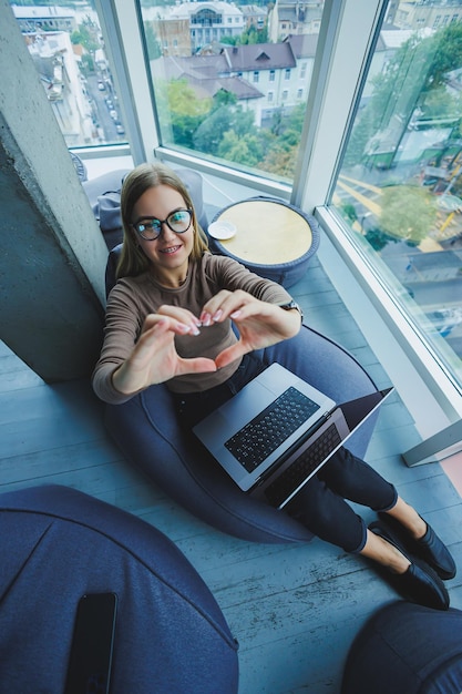 Eine schöne Studentin arbeitet an einem Laptop, während sie auf einem Sitzsack in einem modernen Kreativbüro vor dem Hintergrund von Panoramafenstern sitzt Pausenzeitkonzept