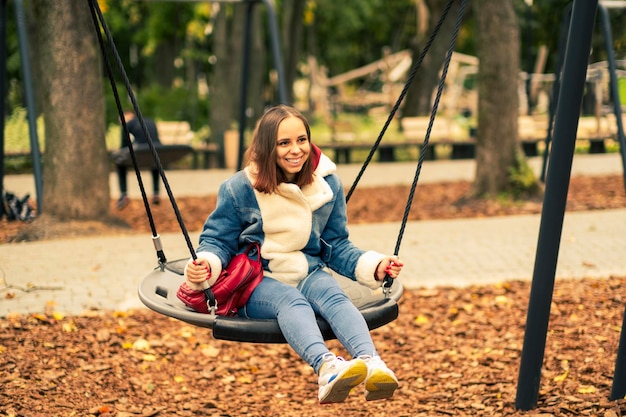 Eine schöne und fröhliche Frau in warmer Kleidung schwingt auf einer Schaukel im Park Mädchen schwingt im Herbststadtpark