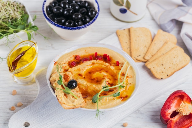 Eine Schüssel Kichererbsen hausgemachter Hummus mit Olivenöl, Knäckebrot, Mikrogrün und geräuchertem Paprika auf weißer Holzoberfläche.