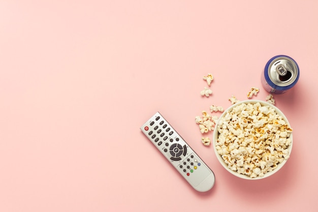 Eine Schüssel Popcorn, eine TV-Fernbedienung, ein Glas Getränk auf einem rosa Hintergrund. Das Konzept, Fernsehen, Film, Fernsehserien, Sport, Shows zu sehen. Flache Lage, Draufsicht.