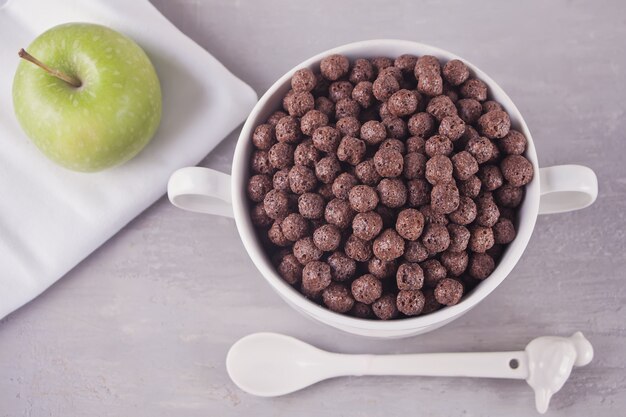 Eine Schüssel trockenes Schokoladenballgetreide und grüner Apfel auf der grauen Tabelle zum Gesundheitsfrühstück