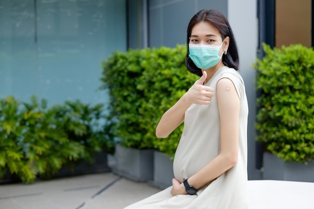 Eine schwangere asiatische Mutter in einem langen Kleid trägt nach der Impfung eine Maske und Daumen hoch.
