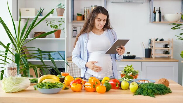Eine schwangere Frau, die ein Tablet verwendet, bereitet gesundes Essen in einer hellen Küche zu