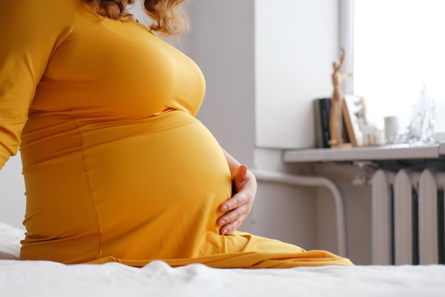 Eine schwangere Frau in einem gelben Kleid sitzt auf dem Bett und streichelt ihren Bauch Nahaufnahme des Bauches