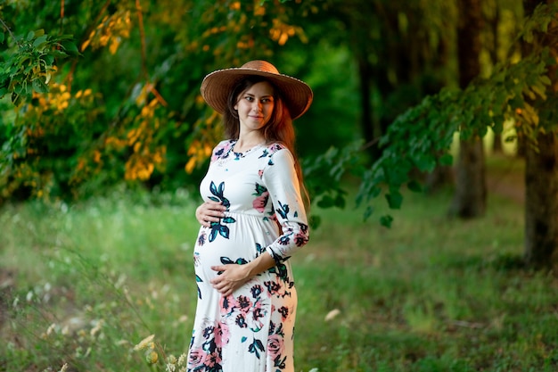 Eine schwangere Frau in einem Kleid und Hut geht im Sommer im Wald spazieren