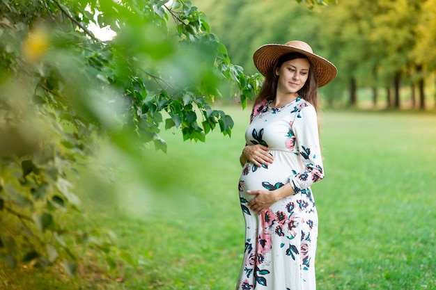 Eine schwangere Frau in einem Kleid und Hut geht im Sommer im Wald spazieren