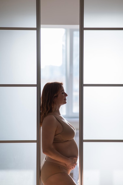 Eine schwangere Frau in nackten Dessous steht im Profil zwischen Falttüren