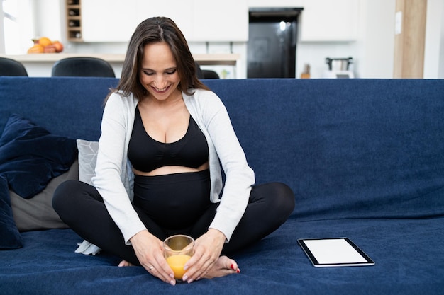 Eine schwangere Frau in Sportkleidung sitzt auf einem blauen Sofa im Wohnzimmer Glas Orangensaft i