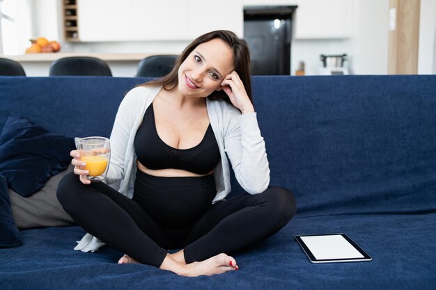 Eine schwangere Frau in Sportkleidung sitzt auf einem blauen Sofa im Wohnzimmer und lächelt in die Kamera