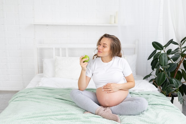 Eine schwangere Frau ist eine junge glückliche werdende Mutter, die einen grünen Apfel isst und zu Hause ihren Bauch auf dem Bett berührt. Das Konzept von Mutterschaft, Schwangerschaft, Kopienraum