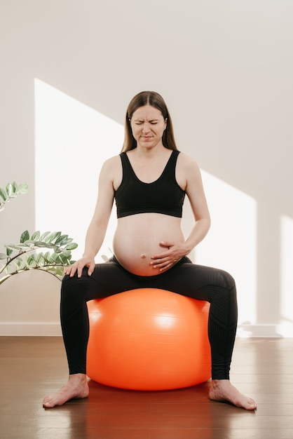 Eine schwangere Frau krümmt sich abends vor Schmerzen mit dem Arm auf dem Bauch auf einem orangefarbenen Gymnastikball bei sich zu Hause. Komplizierte Schwangerschaft.