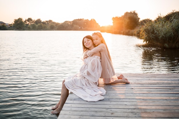 Eine schwangere Frau mit ihrer Tochter vor dem Hintergrund des Wassers bei Sonnenuntergang Eine Frau mit Bauch genießt den Sonnenuntergang am Fluss