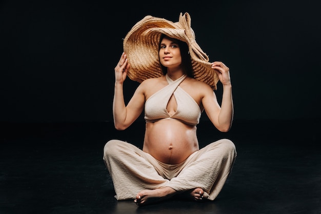Eine schwangere Frau mit Strohhut sitzt in beiger Kleidung in einem Studio auf schwarzem Hintergrund auf dem Boden.