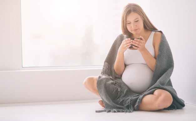 Eine schwangere Frau trinkt Tee oder Kaffee. Ein weibliches Model mit einer Tasse warmem Getränk ist in eine Decke gehüllt