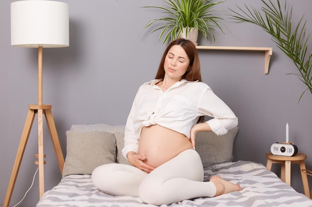 Eine schwangere Frau verspürt Schmerzen im unteren Rücken, die beim Sitzen zu Hause auf dem Bett zu Unwohlsein führen, was möglicherweise auf beginnende Wehen hindeutet