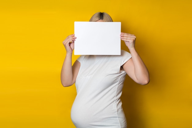 Eine schwangere Frau versteckt ihr Gesicht mit einem sauberen horizontalen Blatt Papier an einer gelben Wand