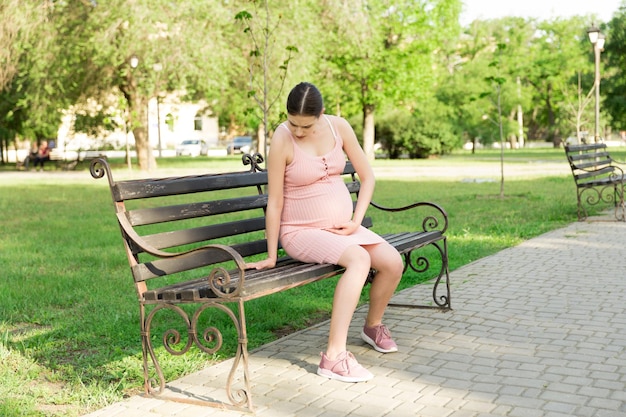 Eine schwangere junge Frau auf einer Bank, die im Park sitzt und sich am Bauch festhält, was Schwangerschaft und Mutterschaft Rückenschmerzen während der Schwangerschaft schmerzt