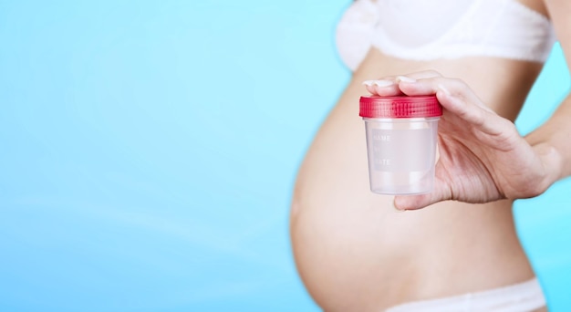 Eine schwangere kaukasische Frau in Unterwäsche, die einen Plastikbehälter zum Sammeln von Urin hält, ein Glas mit einer roten Kappe auf blauem Hintergrund. Nahansicht. Platz kopieren. Foto in hoher Qualität