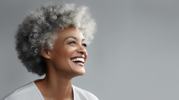 Eine schwarze ältere Frau mit grauen Afrohaaren lächelt und posiert gegen einen hellgrauen Studio-Hintergrund.