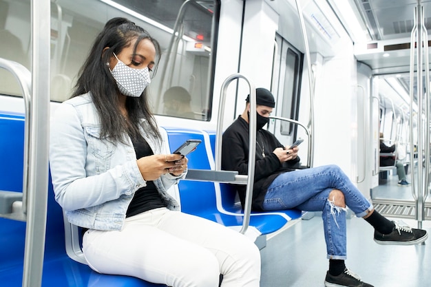 Eine schwarze Frau und ein weißer Mann mit Masken sitzen im U-Bahnwagen unter Verwendung des Smartphones