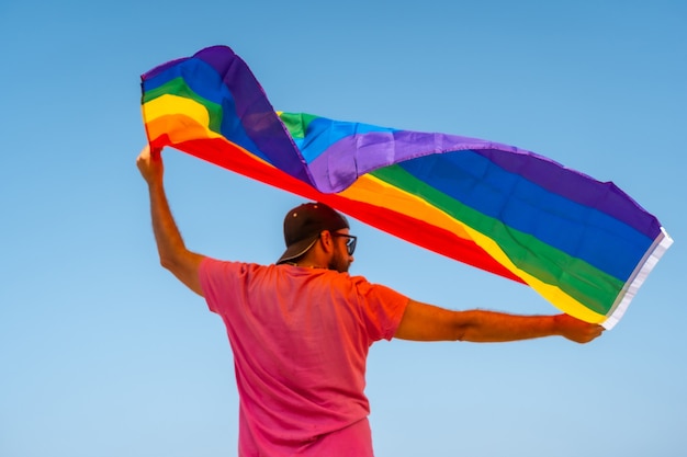 Eine schwule Person mit rosa T-Shirt und schwarzer Mütze mit der LGBT-Flagge auf dem Rücken, die sich im Wind bewegt, mit dem Himmel im Hintergrund, ein Symbol für Homosexualität