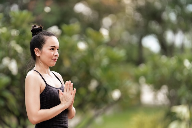 Eine selbstbewusste asiatische Frau mittleren Alters im Sportoutfit, die morgens Yoga-Übungen auf der Yogamatte im Freien im Hinterhof macht. Junge Frau macht Yoga-Übungen im Freien im öffentlichen Naturpark