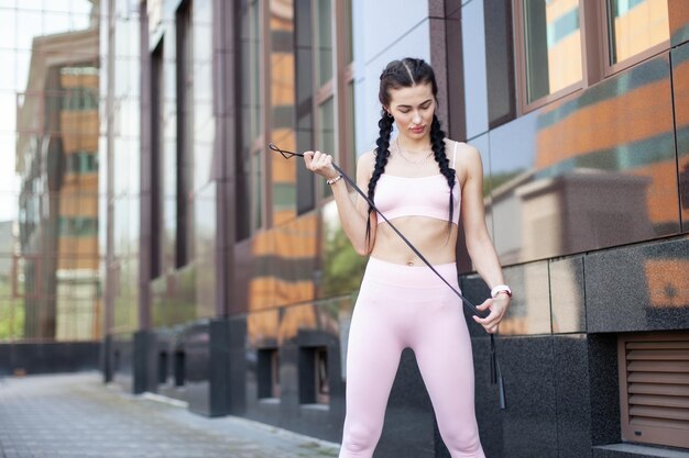 Eine sexy, fitte Frau posiert mit einem Sprungseil in den Händen auf dem Hintergrund eines Stadtgebäudes