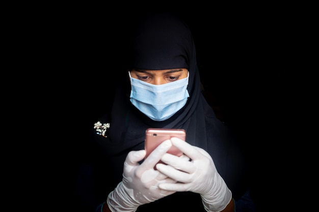 Eine Sicherheitsmaske, die ein asiatisches muslimisches Mädchen trägt, benutzt sein Smartphone Schwarze Hijab-Frau, die eine blaue Maske für die Sicherheit von Coronaviren trägt