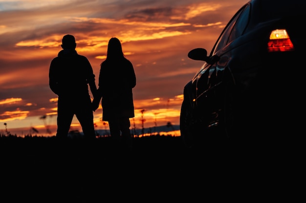 Eine Silhouette eines Paares, das in der Nähe eines Autos vor dem Hintergrund eines schönen Sonnenuntergangs oder Morgenhimmels steht.