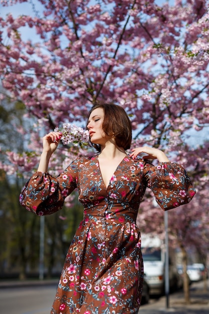 Eine spektakuläre Frau in einem hellen Kleid steht vor dem Hintergrund von Sakura. Eine dunkelhaarige Frau in einem schönen Outfit lächelt beim Gehen auf der Straße