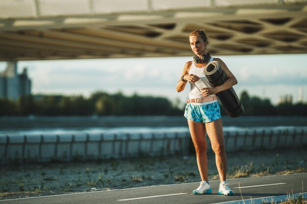 Eine Sportlerin mittleren Alters, die vor dem Training in der Stadt eine Yogamatte und eine Wasserflasche in der Nähe des Flusses hält.