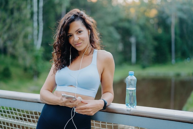 Eine sportliche, brünette junge Frau in Top und Leggings steht auf einer Brücke und hört Musik über Kopfhörer. Sie hält ein modernes Mobiltelefon, trinkt frisches Wasser und trainiert morgens. Menschenmusik-Sportkonzept